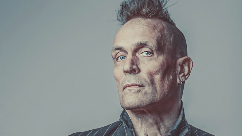 A portrait of a person with a mohawk hairstyle, light blue eyes, and a serious expression, wearing a pinstriped suit jacket and dark shirt, set against a plain grey background. A portrait of a person with a mohawk hairstyle, light blue eyes, and a serious expression, wearing a pinstriped suit jacket and dark shirt, set against a plain grey background.