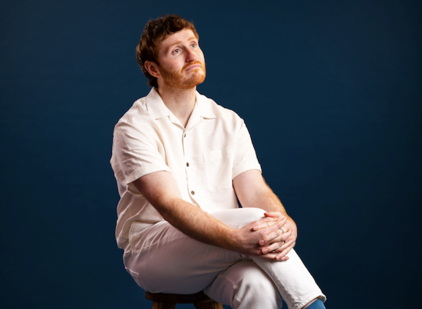 A person wearing white trousers and a white shirt sits on a stool with one leg crossed over. A person wearing white trousers and a white shirt sits on a stool with one leg crossed over.
