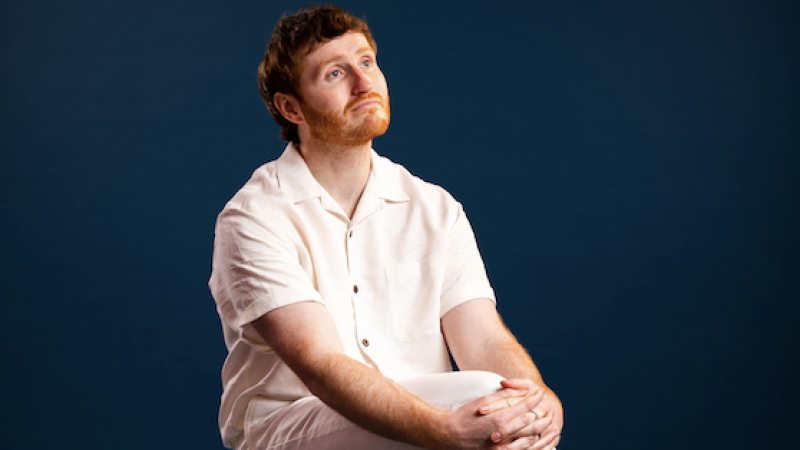 A person wearing white trousers and a white shirt sits on a stool with one leg crossed over. A person wearing white trousers and a white shirt sits on a stool with one leg crossed over.