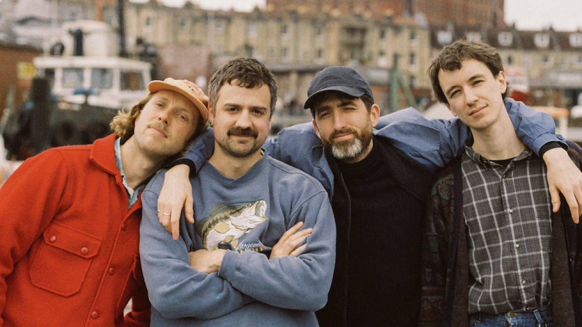Four men standing close together outdoors, smiling and posing for the camera. They appear relaxed and friendly; one is wearing a red jacket and cap, another a blue sweatshirt with a fish print, and the others casual shirts and jackets. A row of old buildings and a boat are visible in the background.