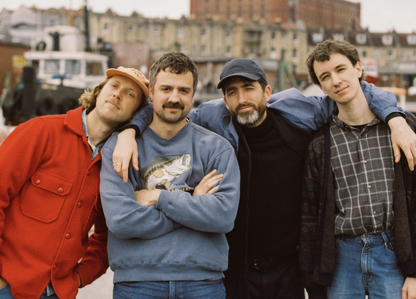 Four men standing close together outdoors, smiling and posing for the camera. They appear relaxed and friendly; one is wearing a red jacket and cap, another a blue sweatshirt with a fish print, and the others casual shirts and jackets. A row of old buildings and a boat are visible in the background.