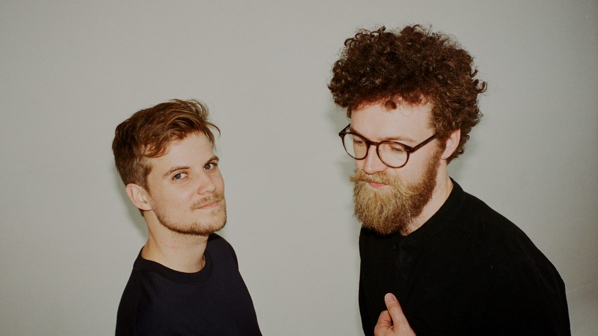 Two men wearing black t shirts stand against a grey background.