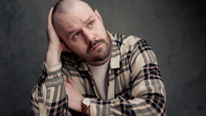 A head and shoulders image of a person leaning on a wooden table. They are in a tan checked shirt, have one hand on their head and are looking off to one side.