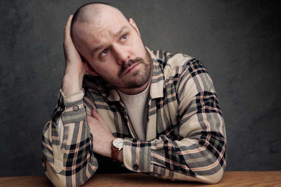 A head and shoulders image of a person leaning on a wooden table. They are in a tan checked shirt, have one hand on their head and are looking off to one side.
