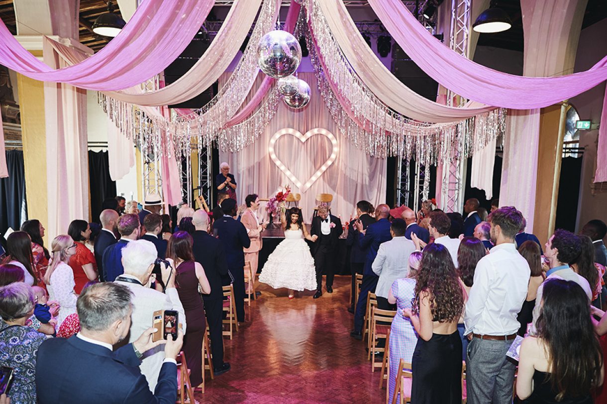 Wedding ceremony at a decorated indoor venue with pink drapes, disco balls, illuminated heart backdrop, and guests seated along the aisle.