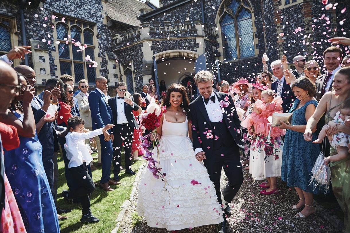 A couple having confetti thrown towards them by a group of guests, in front of a flint building