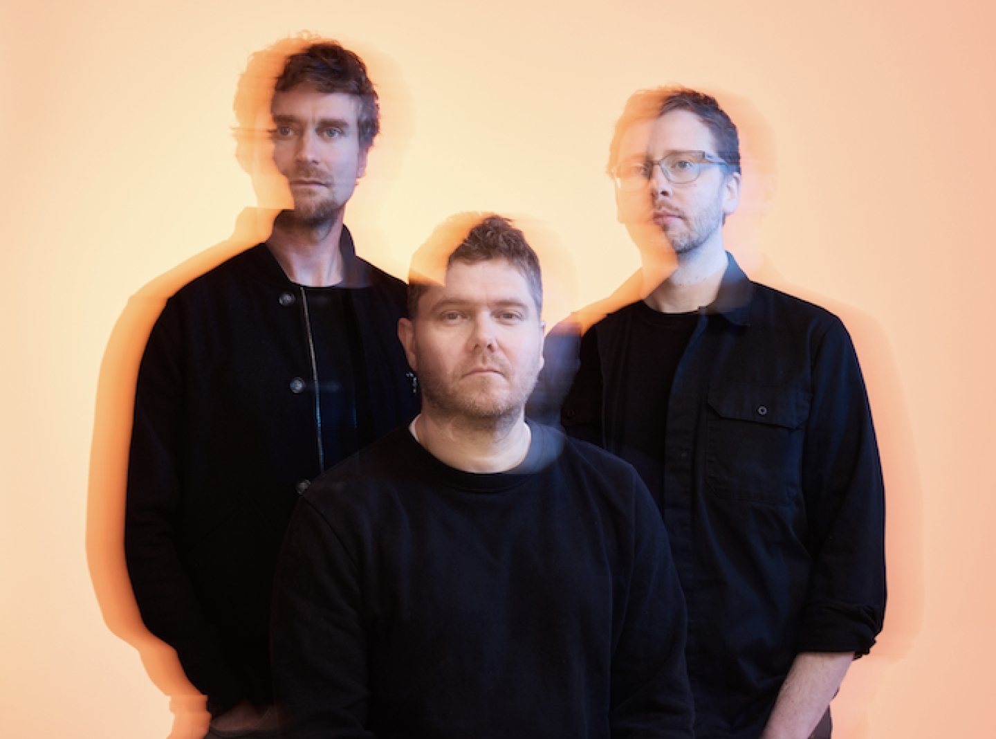 Three people dressed in dark clothing pose against a bright orange background. The image has a motion blur effect, creating double exposures that give a sense of movement or shifting focus.