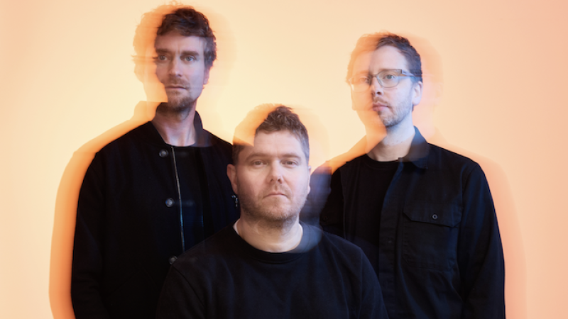 Three people dressed in dark clothing pose against a bright orange background. The image has a motion blur effect, creating double exposures that give a sense of movement or shifting focus.