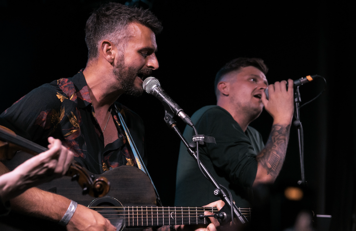 Two men stand against a black background singing into microphones, one is playing the guitar