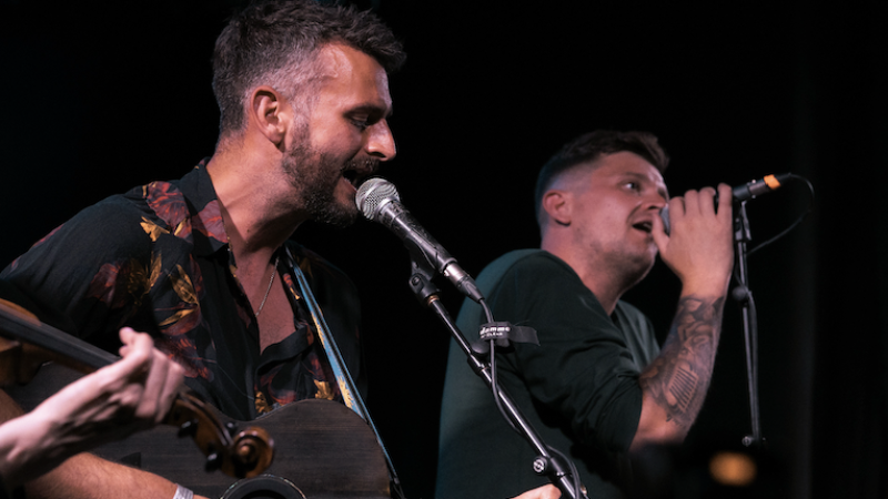 Two men stand against a black background singing into microphones, one is playing the guitar