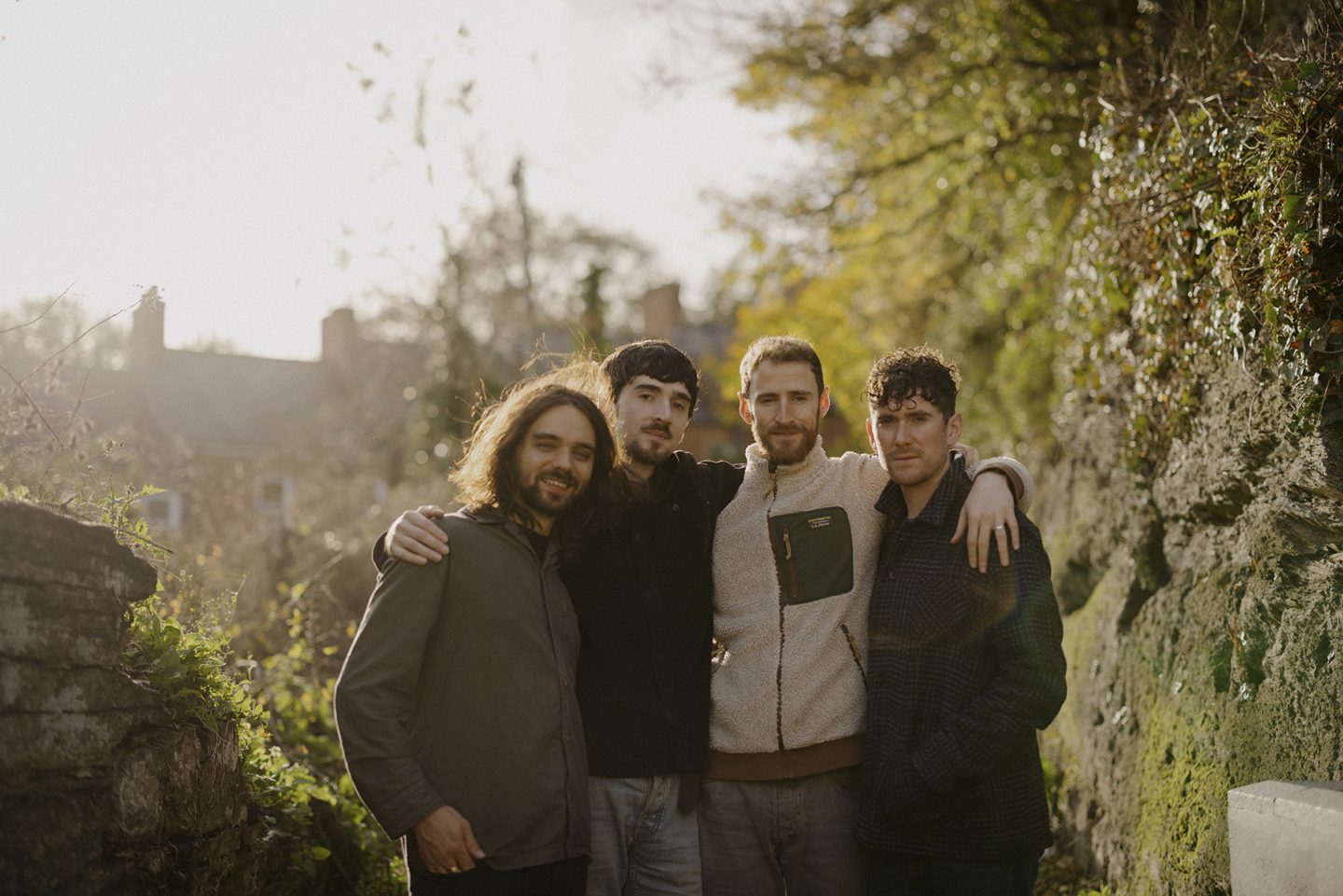 Four men standing close together, with their arms around each other. Behind them is trees and mossy walls.