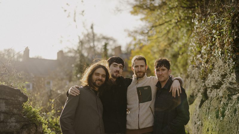 Four men standing close together, with their arms around each other. Behind them is trees and mossy walls.