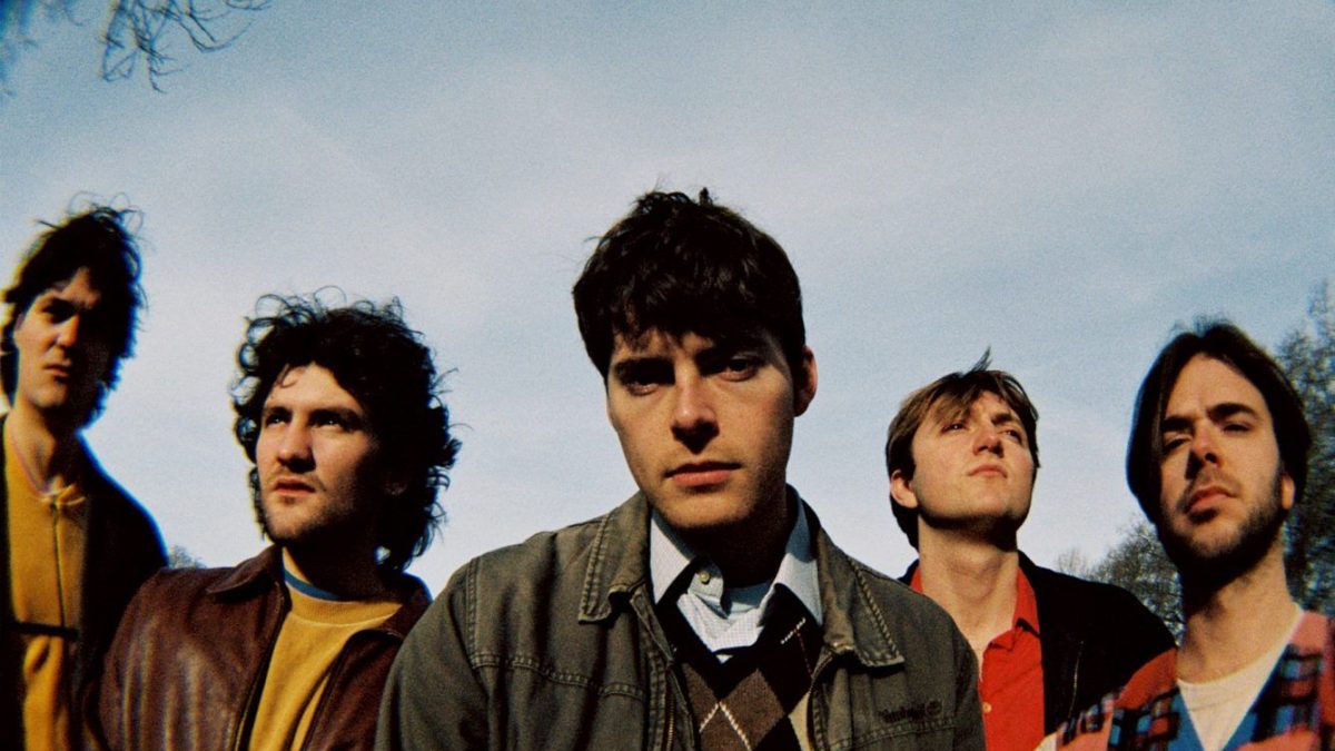 A head and shoulders image of 5 people with short dark hair stand close together, with a sky behind them.