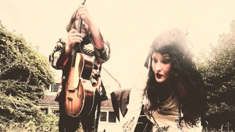 Two people outdoors near a house, one holding an acoustic guitar and the other leaning forward, surrounded by greenery.