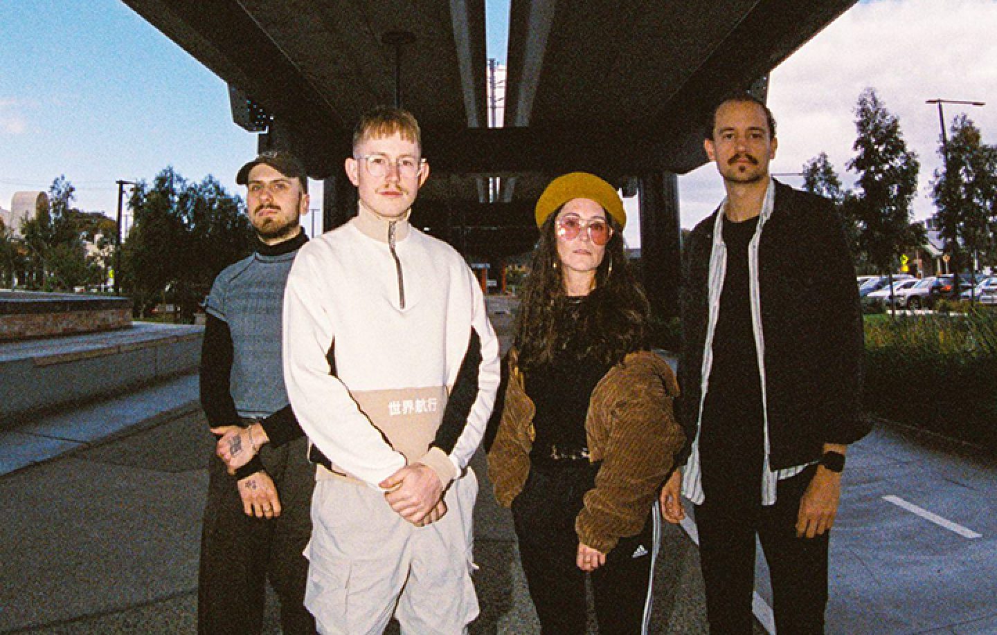 The image shows four individuals standing outdoors beneath an elevated structure, likely a bridge or overpass. Parked cars are visible on the right side, and the sky is partly cloudy with patches of blue.