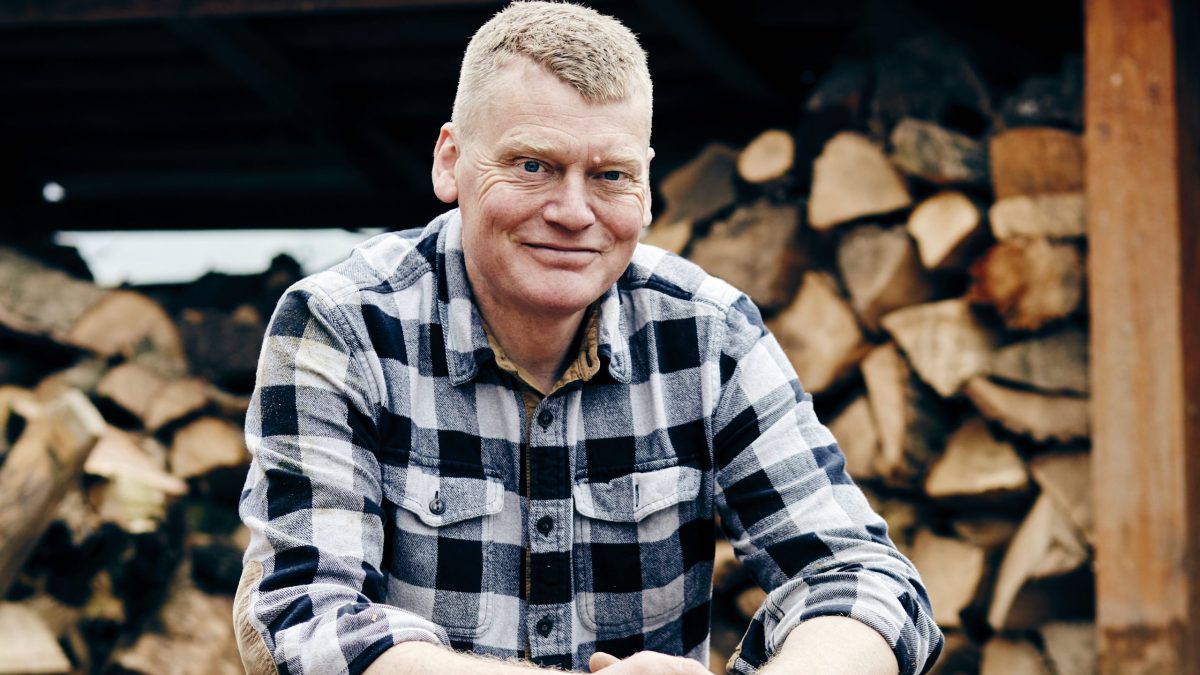 Person wearing a checkered flannel shirt sitting in front of a large stack of chopped firewood.