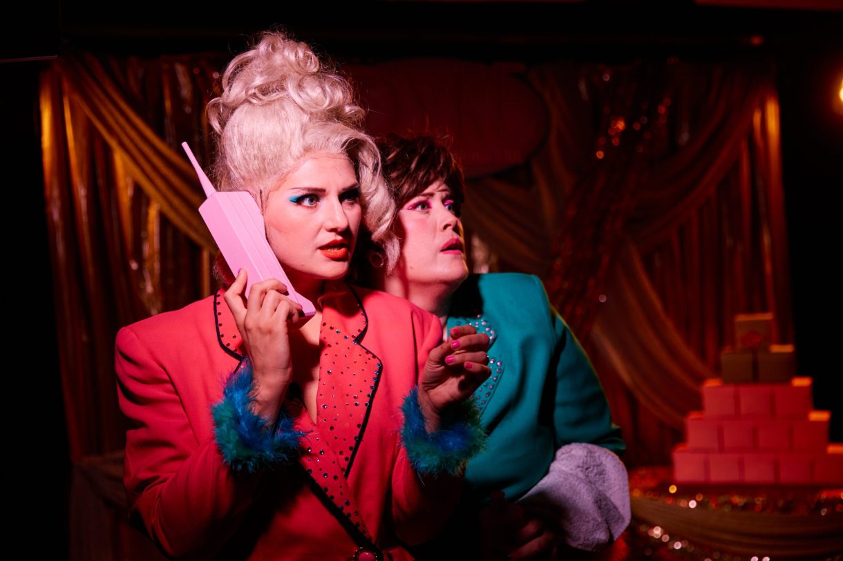 Two people in bright retro outfits under dramatic lighting. One wears a pink jacket with blue feathered cuffs and holds a pink novelty phone. The other wears a teal jacket with pink feathered cuffs. Gold draped fabric and stacked pink boxes form the background.