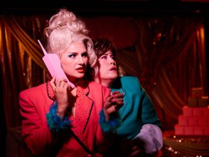 Two people in bright retro outfits under dramatic lighting. One wears a pink jacket with blue feathered cuffs and holds a pink novelty phone. The other wears a teal jacket with pink feathered cuffs. Gold draped fabric and stacked pink boxes form the background.
