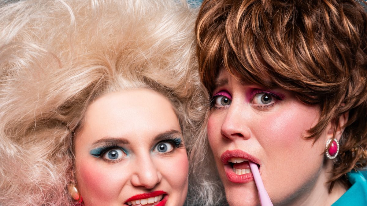 Two people in bold retro glam outfits pose together, one holding cash and the other a pink cordless phone.