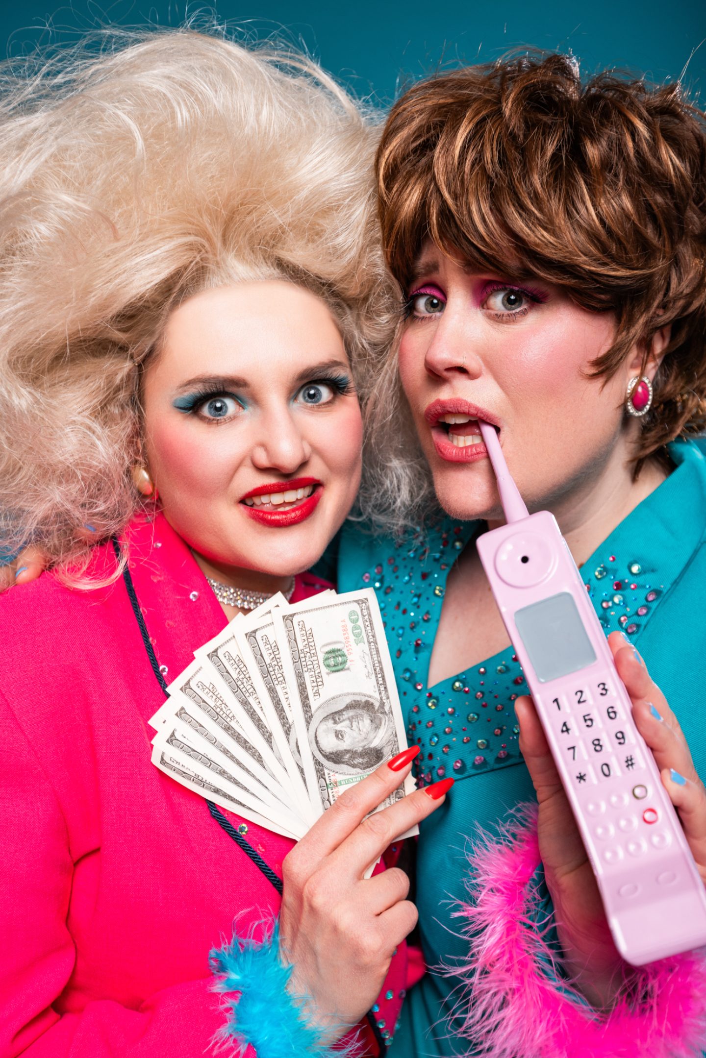 Two people in bold retro glam outfits pose together, one holding cash and the other a pink cordless phone.