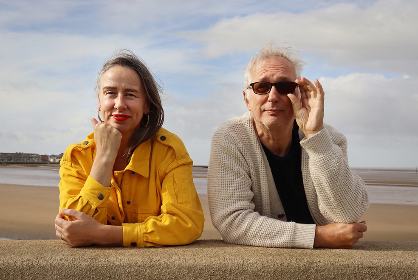 The image shows two individuals leaning on a stone or concrete barrier along a beach promenade. The person on the left is wearing a bright yellow button-up jacket, while the person on the right is dressed in a light-colored knitted cardigan over a dark shirt. Behind them is a sandy beach with shallow water and a distant shoreline.