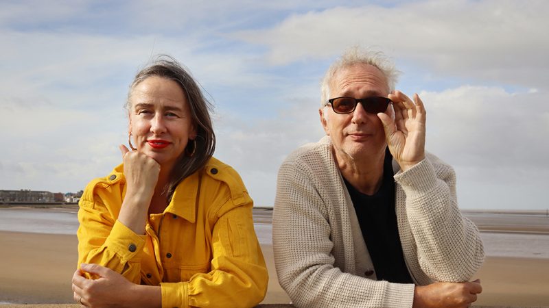 The image shows two individuals leaning on a stone or concrete barrier along a beach promenade. The person on the left is wearing a bright yellow button-up jacket, while the person on the right is dressed in a light-colored knitted cardigan over a dark shirt. Behind them is a sandy beach with shallow water and a distant shoreline.