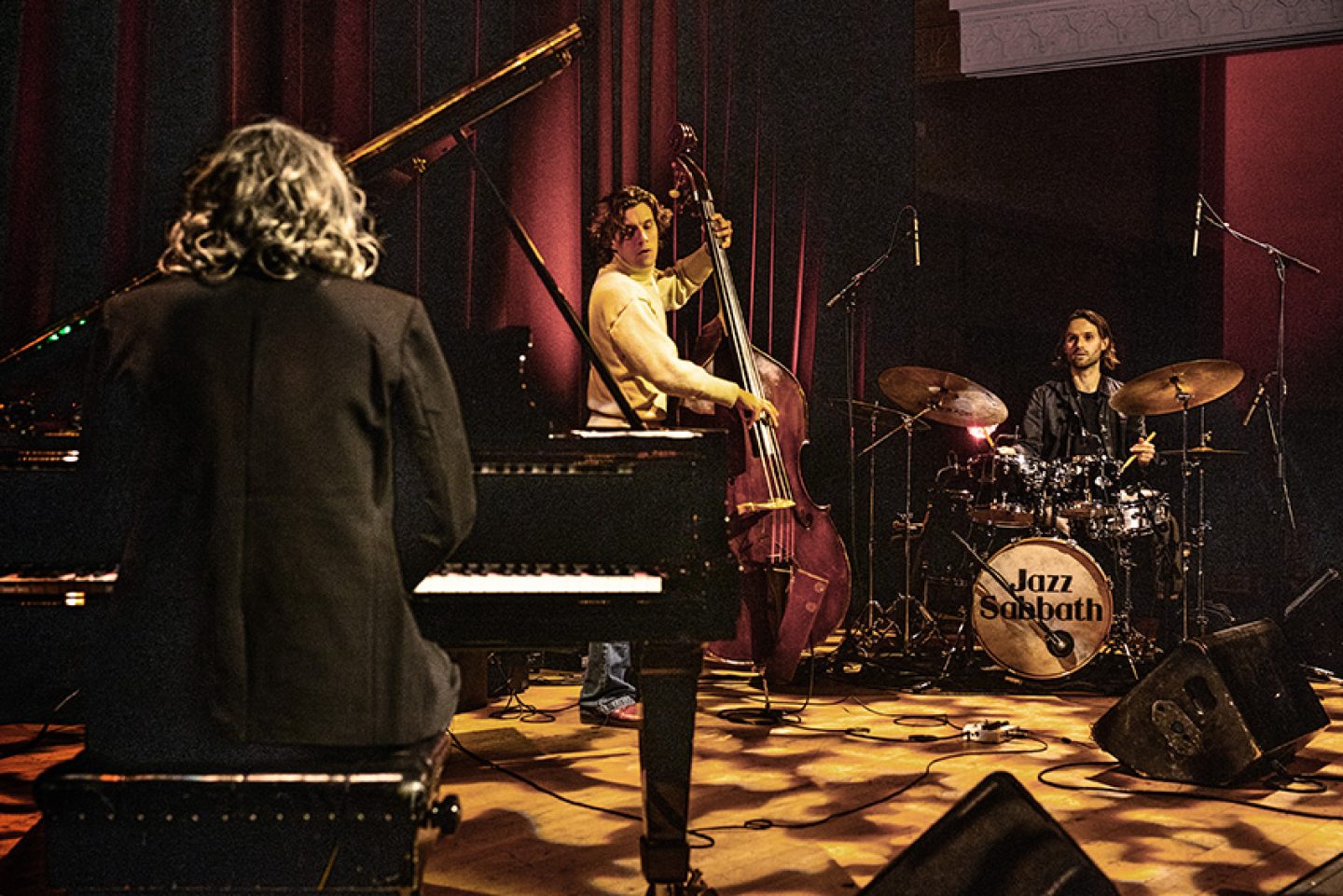 The image shows a live jazz performance on a stage with three musicians playing different instruments. In the foreground, a musician with curly hair is seated at a grand piano, facing away from the camera. To the left, another musician is standing and playing an upright double bass. On the right side of the stage, a drummer is positioned behind a drum kit, which prominently displays the words “Jazz Sabbath” on the bass drum.