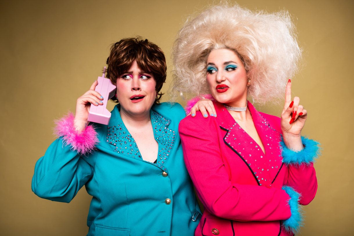 Two people in bold, retro outfits against a mustard background. One wears a teal jacket with pink feathered cuffs and holds a pink novelty phone. The other wears a bright pink jacket with blue feathered cuffs and has large, voluminous platinum-blonde hair.