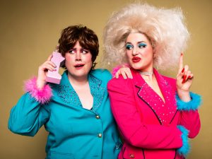 Two people in bold, retro outfits against a mustard background. One wears a teal jacket with pink feathered cuffs and holds a pink novelty phone. The other wears a bright pink jacket with blue feathered cuffs and has large, voluminous platinum-blonde hair.