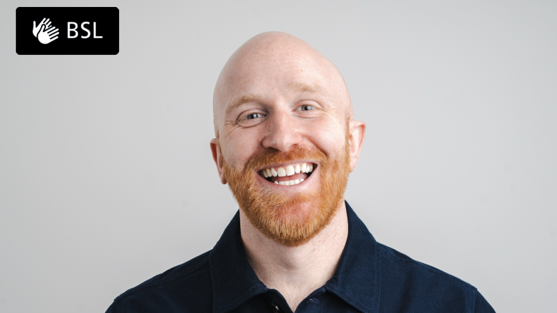A head and shoulder image of a person looking at the camera. They have a full, orange beard and is smiling widely. They are wearing a dark blue collared shirt. Logo on image reads BSL.