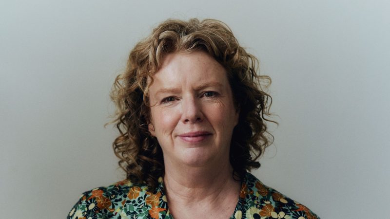 Person with curly hair wearing a patterned shirt featuring green, orange, and white floral designs, standing against a plain light-colored background.