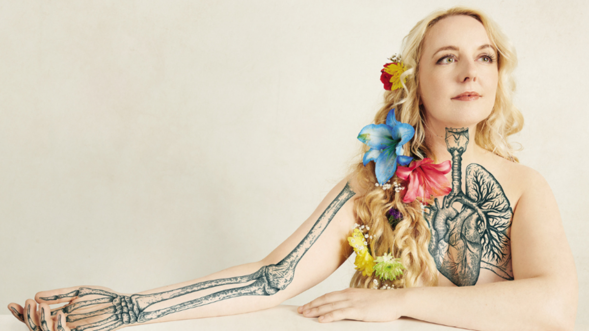 Person with long, wavy hair adorned with colorful flowers, sitting against a light background. Visible body art includes a detailed anatomical heart on the chest, a skeletal arm design extending from the shoulder to the hand, and a neck tattoo connecting the two.