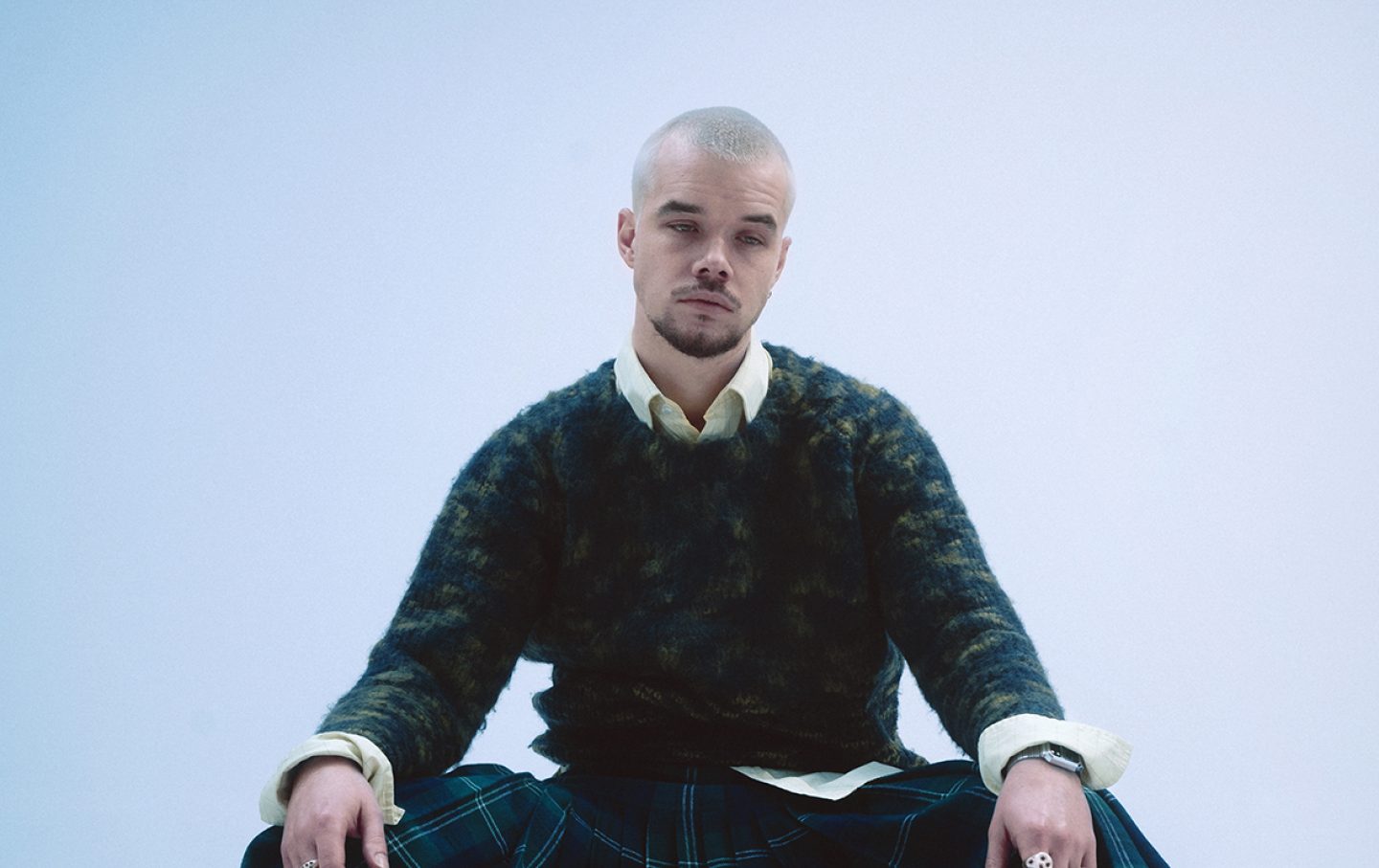 A person on a white background, sat with their hands wrapped over their knees. They have short bleached hair and a black jumper over a yellow shirt.