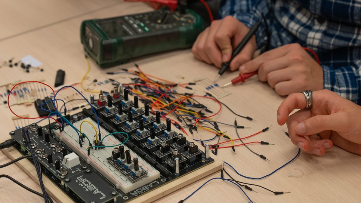 Electronics workshop table with a circuit board featuring multiple knobs and connectors, surrounded by colorful wires, resistors, and small components. Two hands work on the setup with a probe, and a green multimeter is in the background.