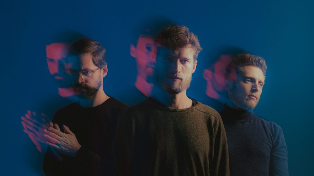 Three men against a blue background, shown with layered red and blue motion blur that creates ghosted, overlapping silhouettes and a moody, introspective feel.