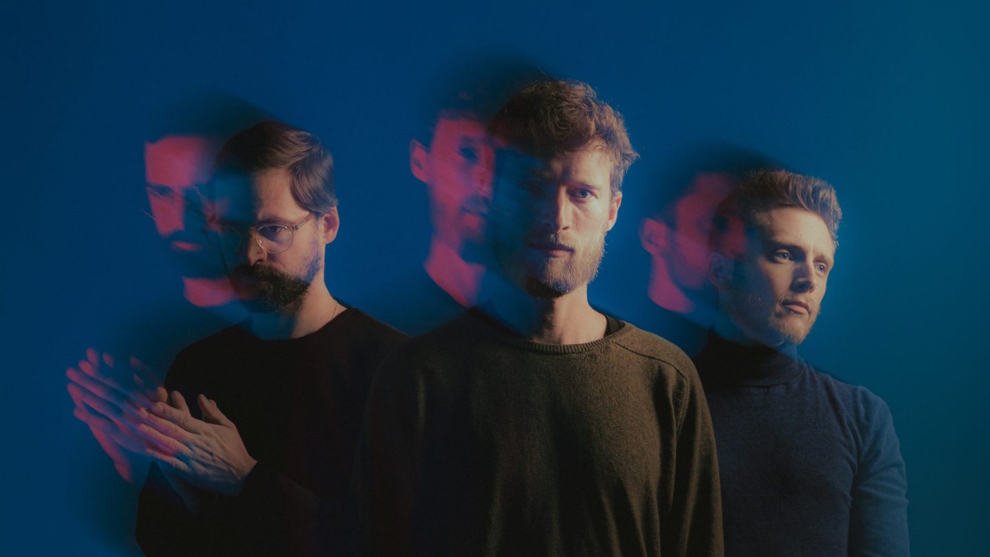 Three men against a blue background, shown with layered red and blue motion blur that creates ghosted, overlapping silhouettes and a moody, introspective feel.