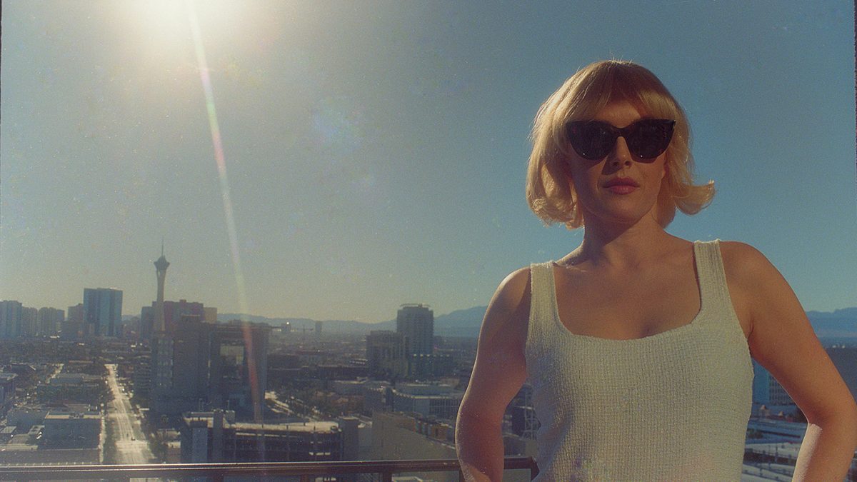 A woman in sunglasses stands on a balcony overlooking a sunlit city skyline.