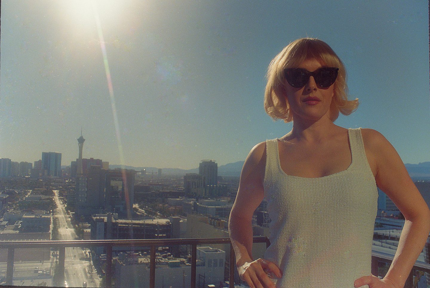 A woman in sunglasses stands on a balcony overlooking a sunlit city skyline.