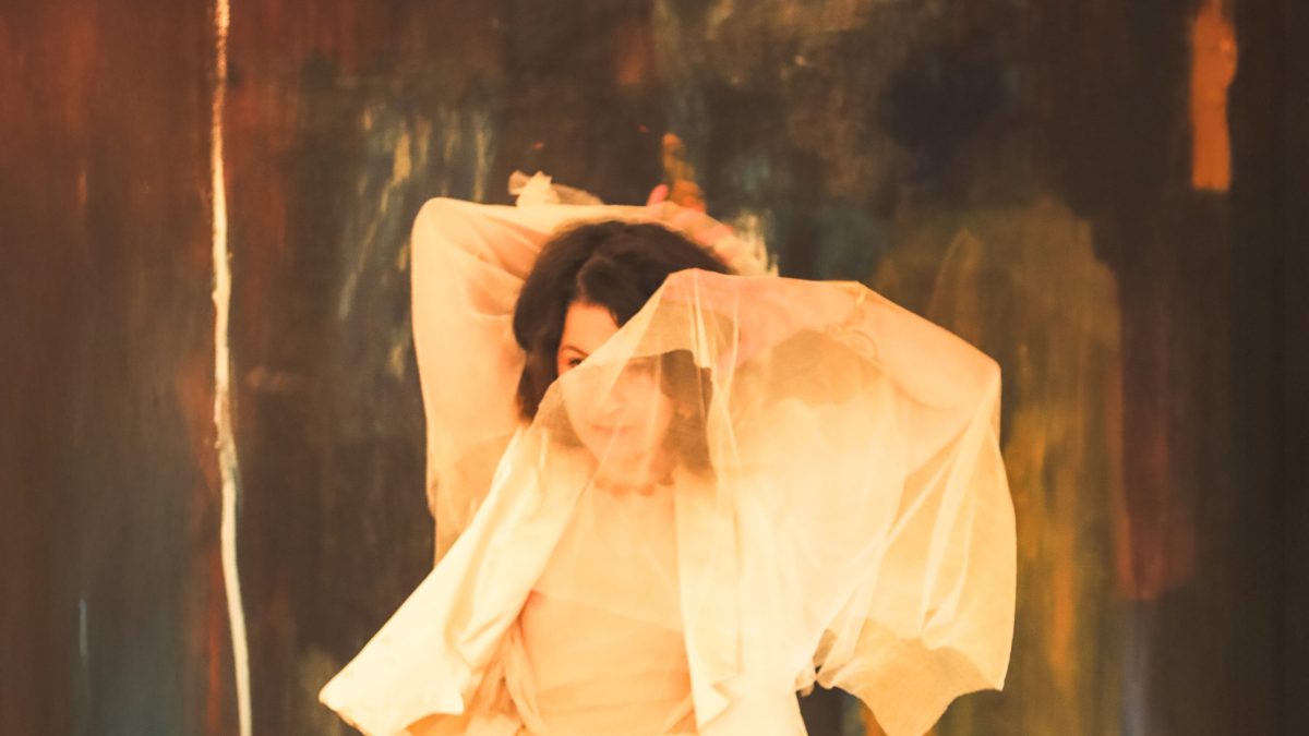 Person in flowing light fabric with arms raised, set against an abstract painted background.
