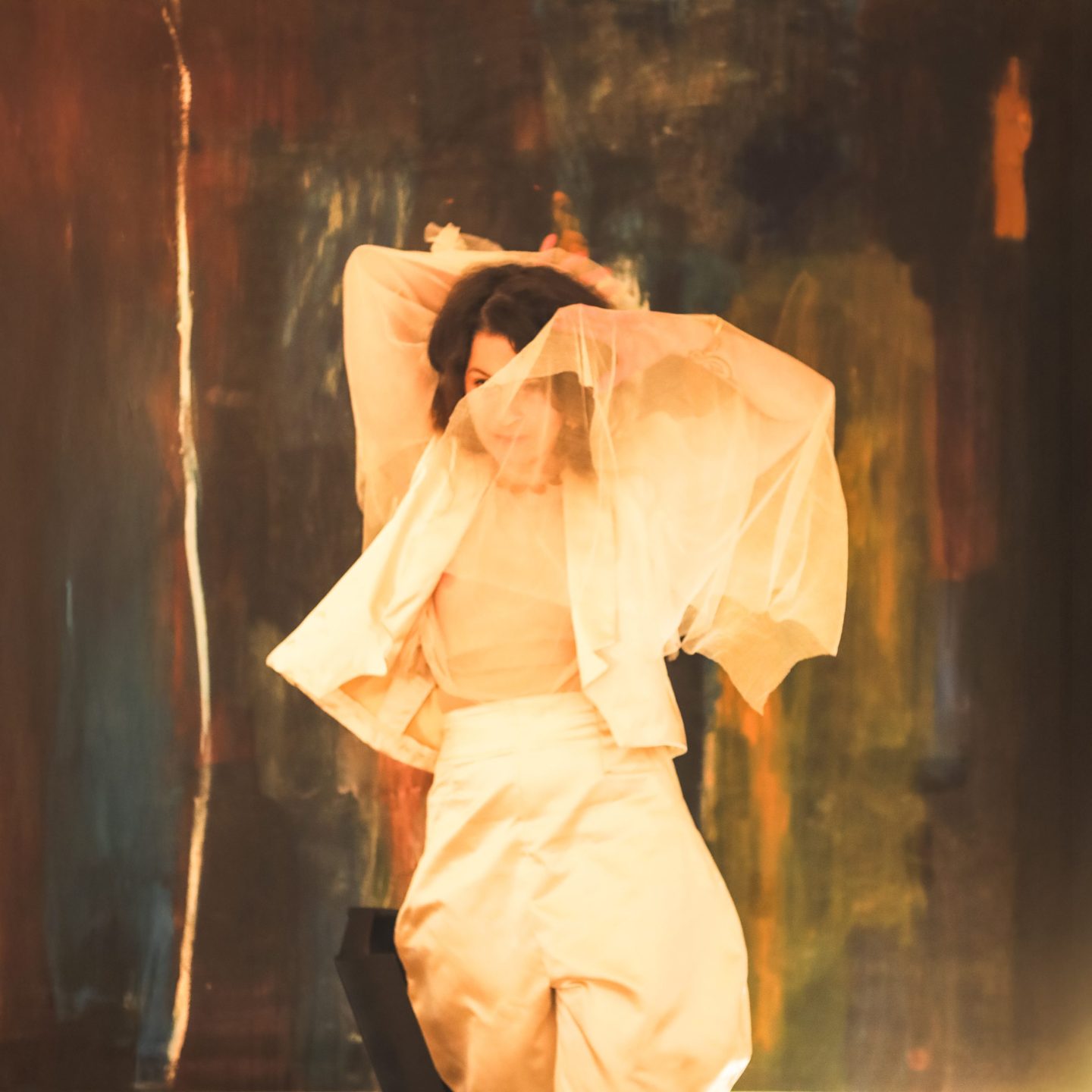 Person in flowing light fabric with arms raised, set against an abstract painted background.