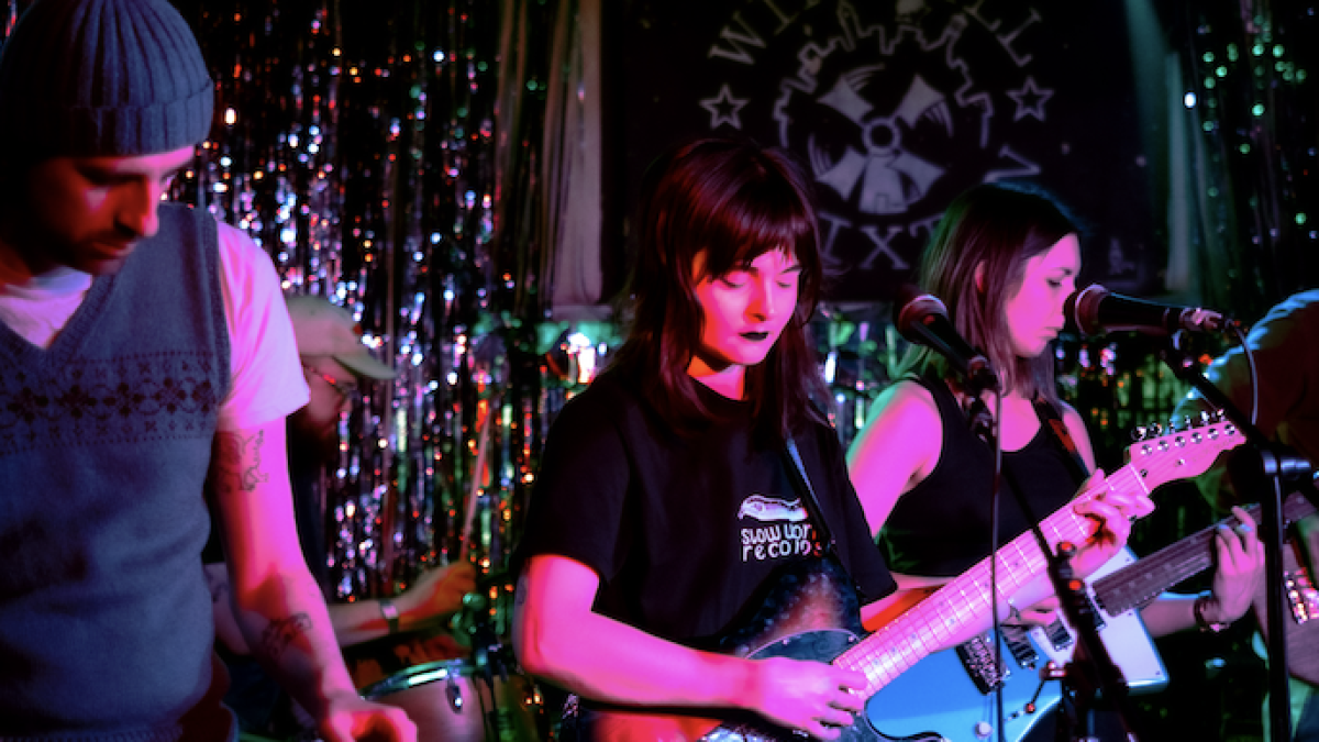 Three musicians play guitars and a keyboard on a small, colorful, dimly lit stage.