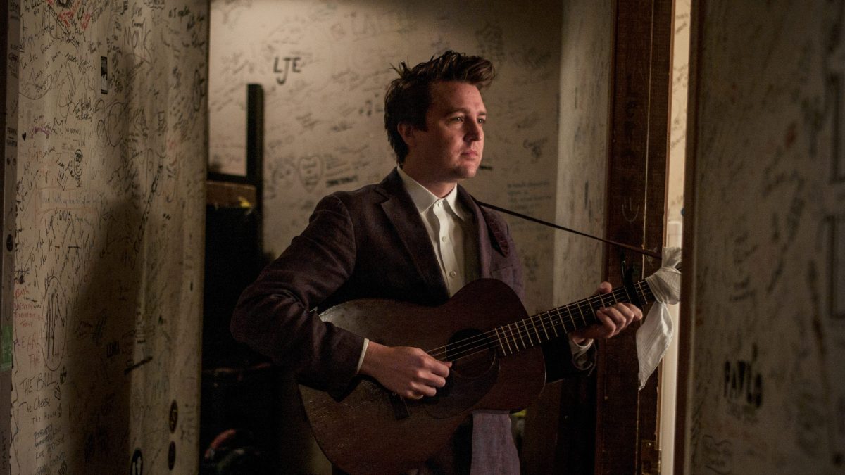 Musician holding an acoustic guitar in a graffiti-covered backstage hallway.