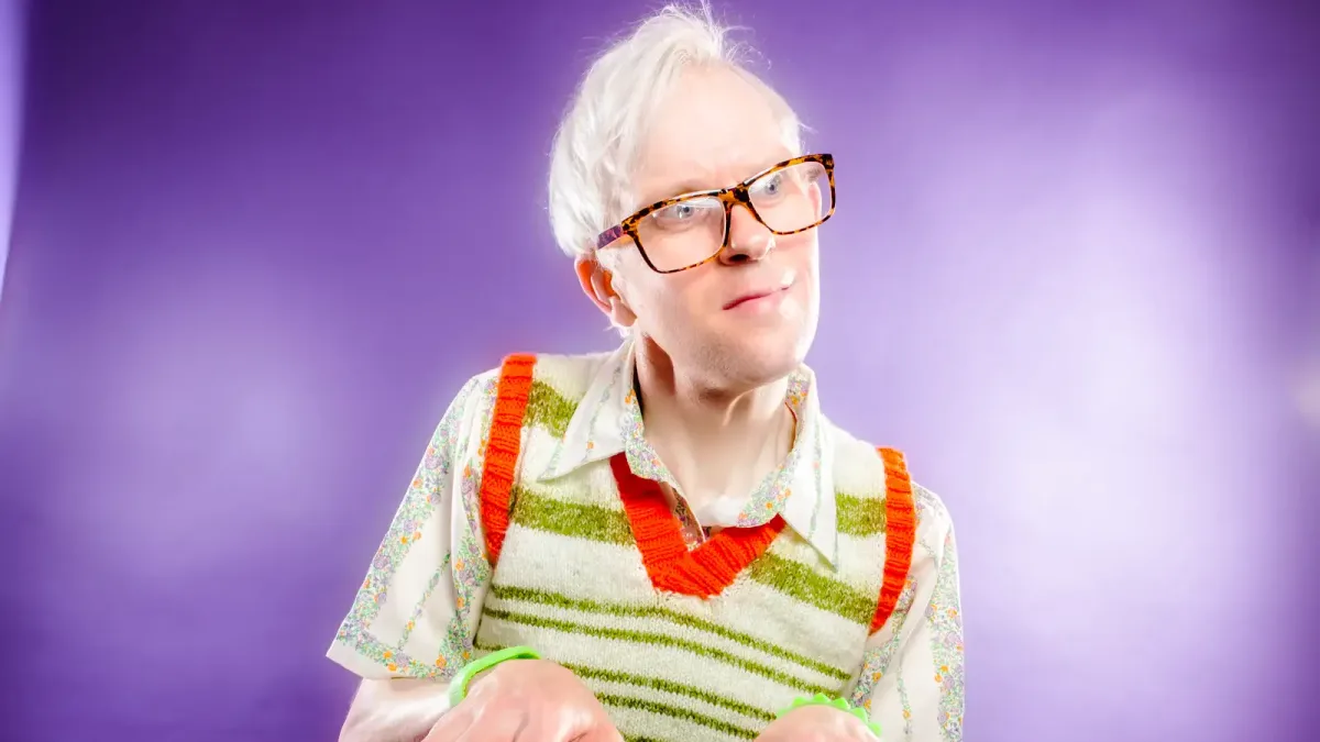 A white-haired person wearing large tortoiseshell glasses and a striped knit sweater vest plays an electronic keyboard against a purple background, tilting their head with a curious expression.