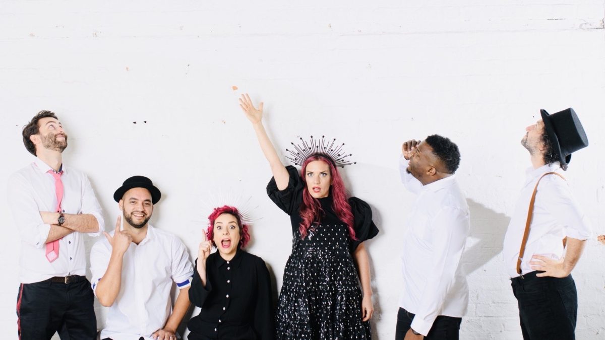 A group of six people pose playfully against a white brick wall. They are dressed in a mix of black-and-white outfits with creative, quirky styling. One person in the center stands with an arm raised and wears a black dress adorned with sparkles and a spiked crown-like headpiece. Two people are seated in front, one wearing striped tights and the other in a white shirt with a hat. The remaining three stand around them, each striking expressive or whimsical poses. The scene has a fun, energetic, and theatrical atmosphere.