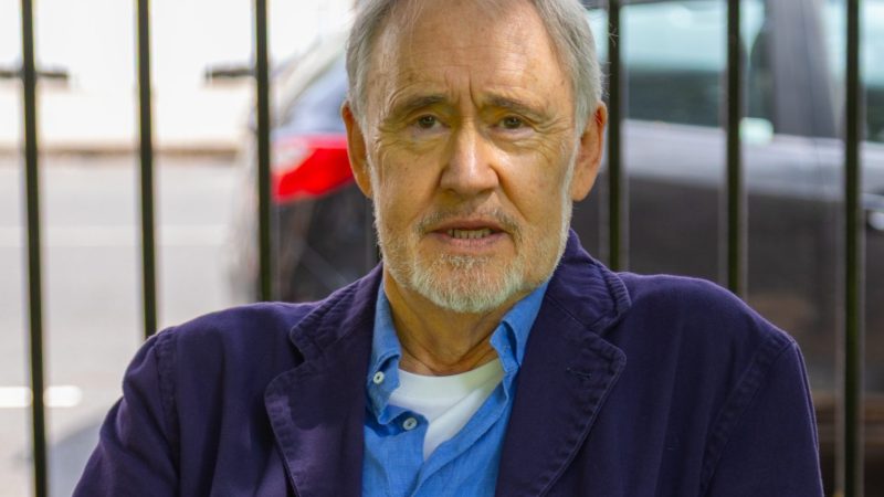 An older man sits on a bench outdoors, wearing a blue jacket and shirt, holding an orange lanyard, with a calm, thoughtful expression.