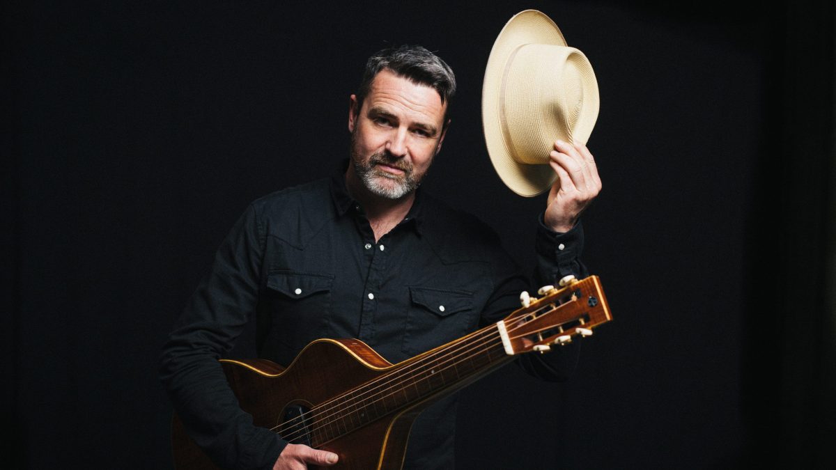 A person dressed in a dark button-up shirt holds an acoustic guitar across their body while raising a light-colored cowboy-style hat in one hand. They stand against a dark background.