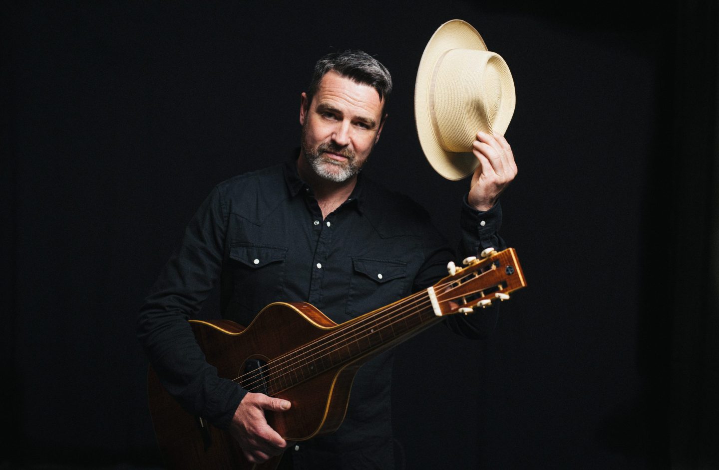 A person dressed in a dark button-up shirt holds an acoustic guitar across their body while raising a light-colored cowboy-style hat in one hand. They stand against a dark background.