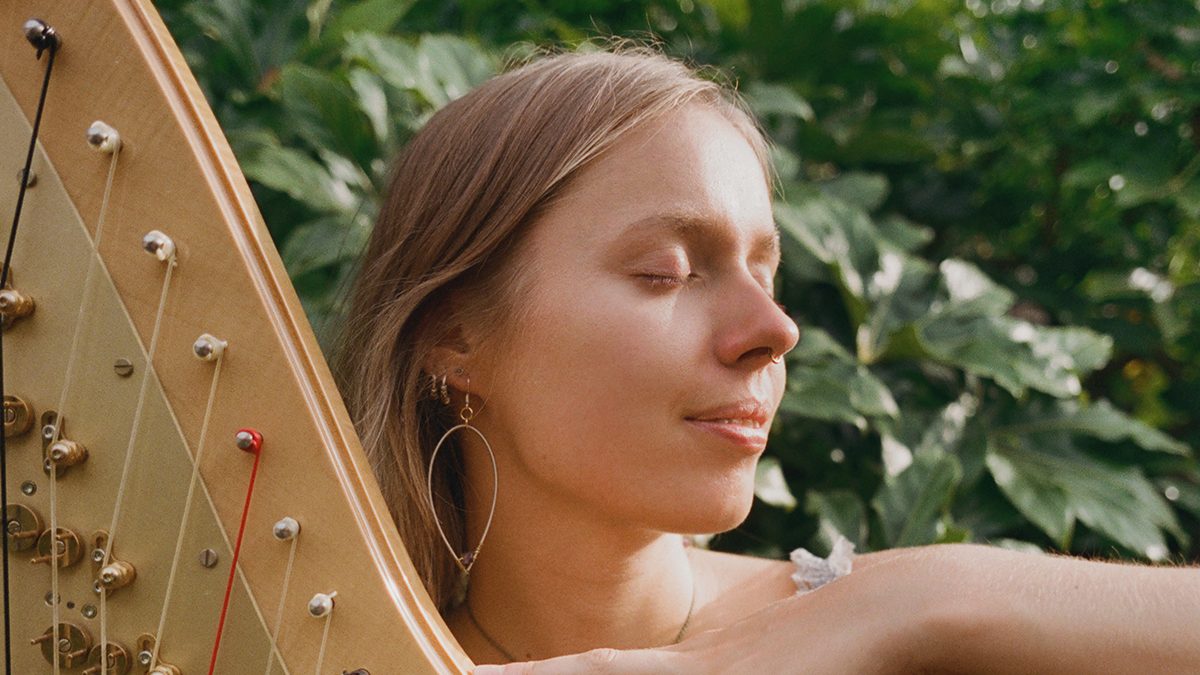 A person outdoors holding a wooden harp, with one hand resting on the instrument’s strings. The harp has a curved frame with multiple tuning pins and strings in various colors. Lush green foliage fills the background.