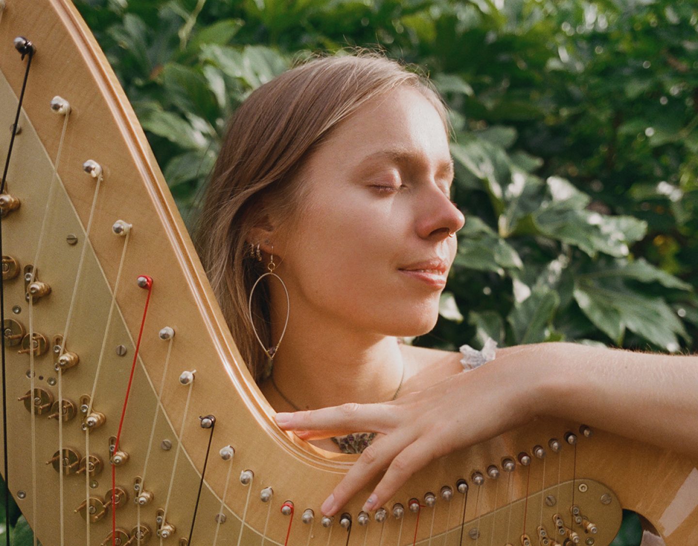 A person outdoors holding a wooden harp, with one hand resting on the instrument’s strings. The harp has a curved frame with multiple tuning pins and strings in various colors. Lush green foliage fills the background.
