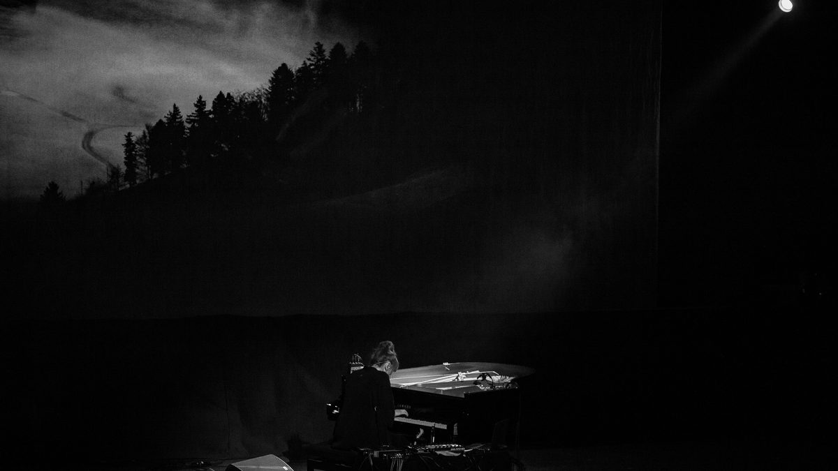 A black‑and‑white photograph of a musician seated at a piano on a dimly lit stage. The performer is shown from behind, illuminated by a soft overhead light. Cables and equipment surround the piano. In the background, a large screen displays a dark, atmospheric image of a forested hillside under cloudy skies, creating a moody and cinematic setting.